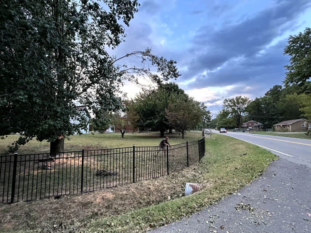 A two-lane road with a nearby fenced area, trees, and houses is depicted under a cloudy sky.